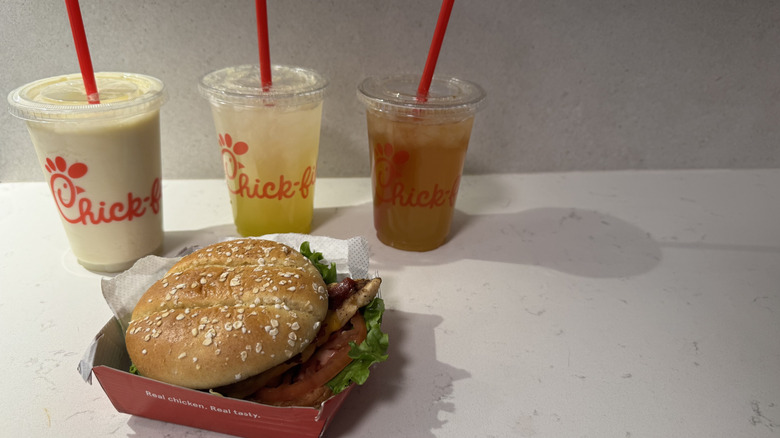 Three Chick-fil-A drink cups with a chicken sandwich on white counter.