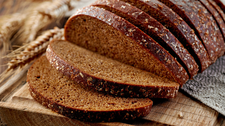 Dark rye bread loaf, sliced on a cutting board with fresh rye in the background