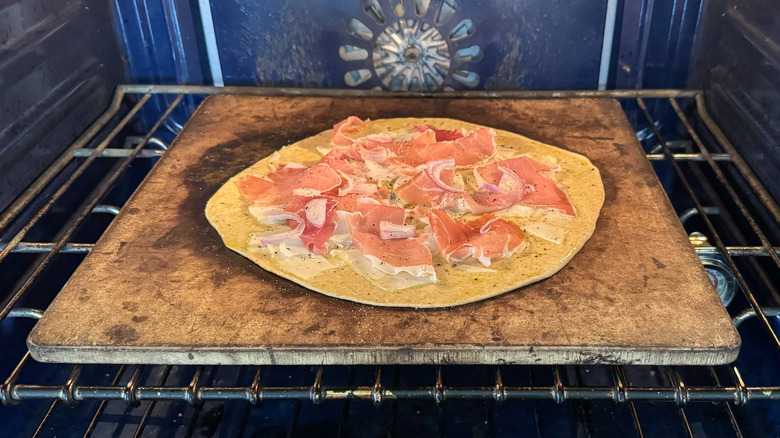 Flatbread with shaved goat gouda, torn prosciutto, sliced shallots, and black pepper on pizza stone in oven