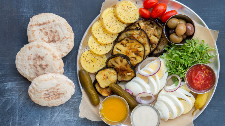 Sabich salad with eggplant, eggs, potatoes, olives, pickles, greens, and sauces on metal plate with pita bread