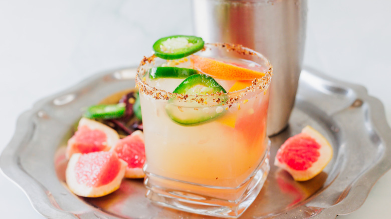 A glass of margarita with garnishes and shaker on metal tray