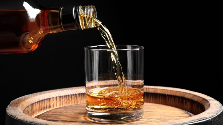 Bourbon being poured from a bottle into a glass on an oak barrel