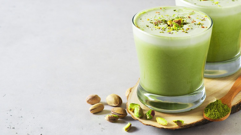 two pistachio lattes on wooden serving platter with pistachio nuts and spoon of matcha powder