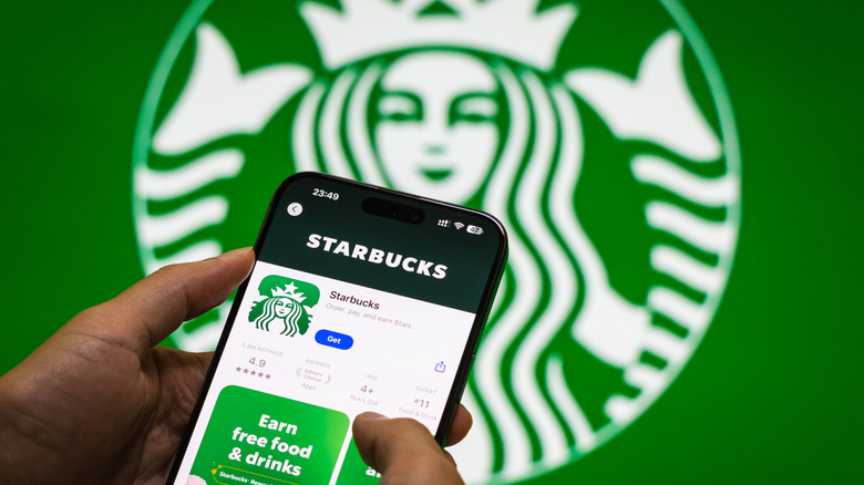 Hand holding a smartphone with the Starbucks mobile app in front of a big Starbucks logo