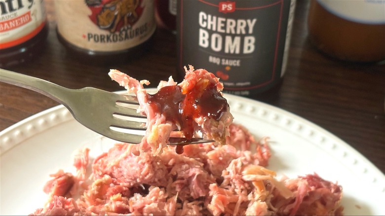 Fork with pulled pork and BBQ sauce above plate of pulled pork and bottles of BBQ sauces