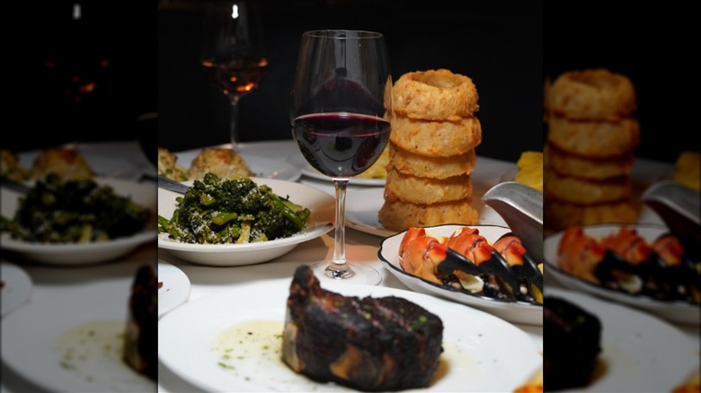 Steak, broccoli, stone crabs, and extra-large onion rings surrounding wine glass on white cloth table