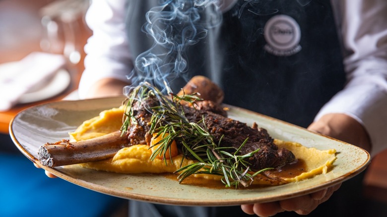 Server holds steaming plate of meat with polenta and sprig of rosemary