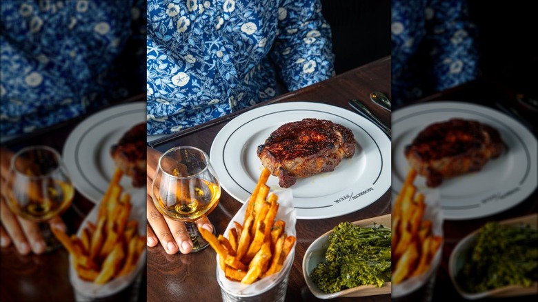 Man in blue floral shirt sitting at table with steak and side of fries, broccolini, and white wine