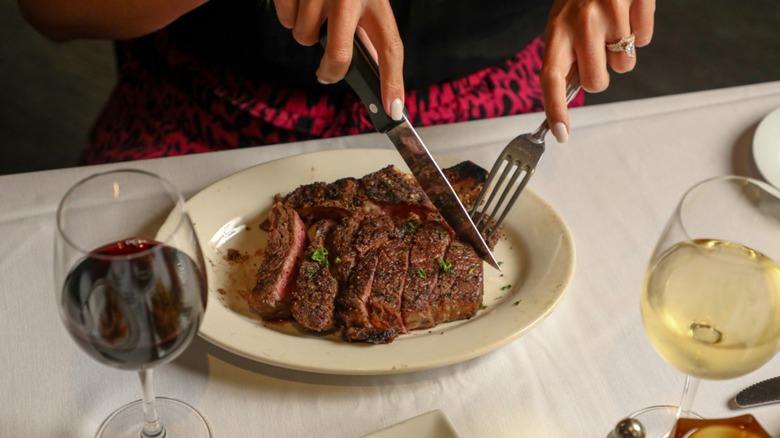 Woman cuts steak next to glass of red wine on white cloth table