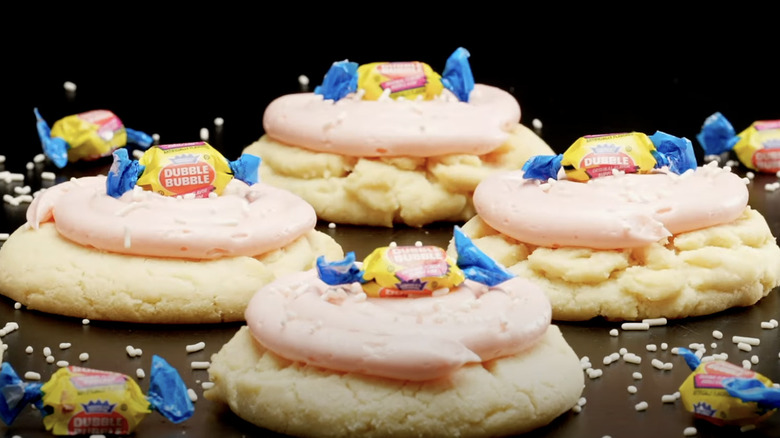 4 sugar cookies with pink frosting and white background with a wrapped piece of bubble gum on top of each