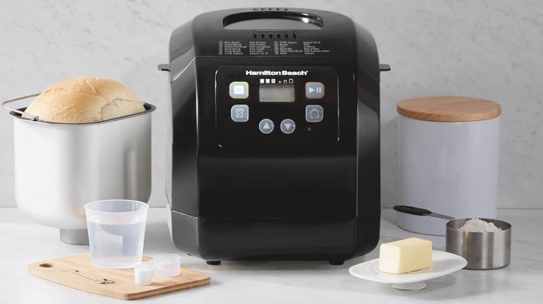 Hamilton Beach bread maker in black sitting on counter next to accessories, fresh bread, and butter