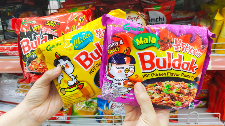 Hands holding three Buldak ramen packages in a store aisle