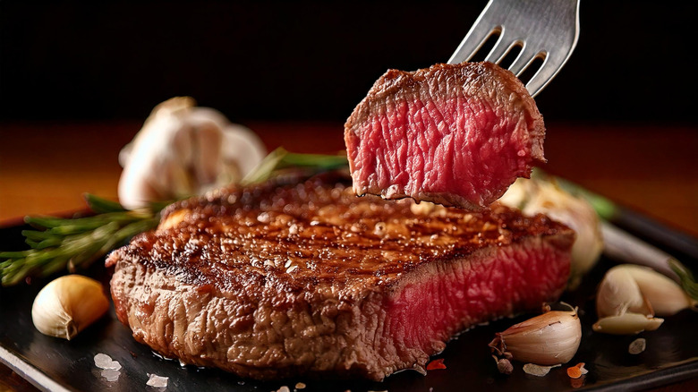Fork lifting slice of steak out of filet surrounded by garlic cloves and rosemary