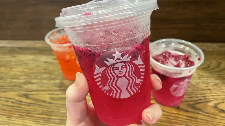 a person holding a cup of mango dragonfruit lemonade refresher in a classic Starbucks plastic cup with other refreshers in the back