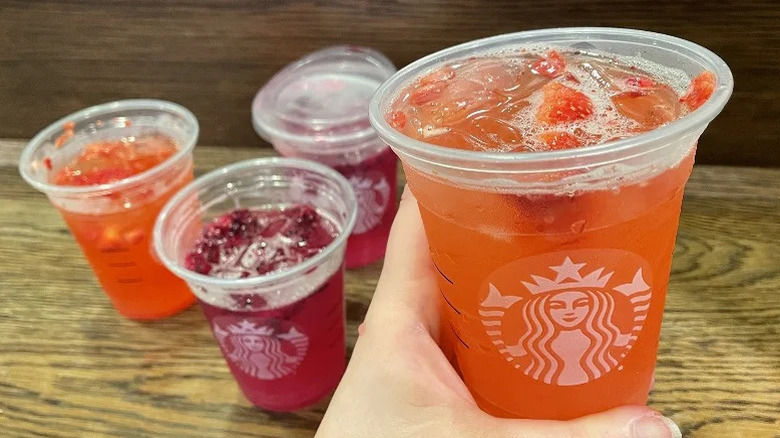 a person holding a strawberry acai lemonade refresher from starbucks with other drinks in the back