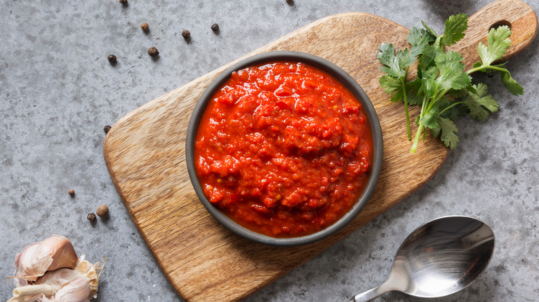 bowl of ajvar atop a wooden board with garlic, pepper, and herbs