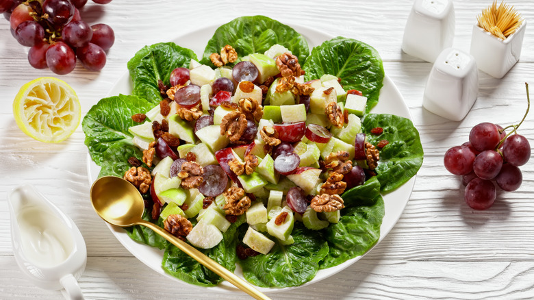 A Waldorf salad served on a white plate, on a table surrounded by lemons, grapes, dressing and more