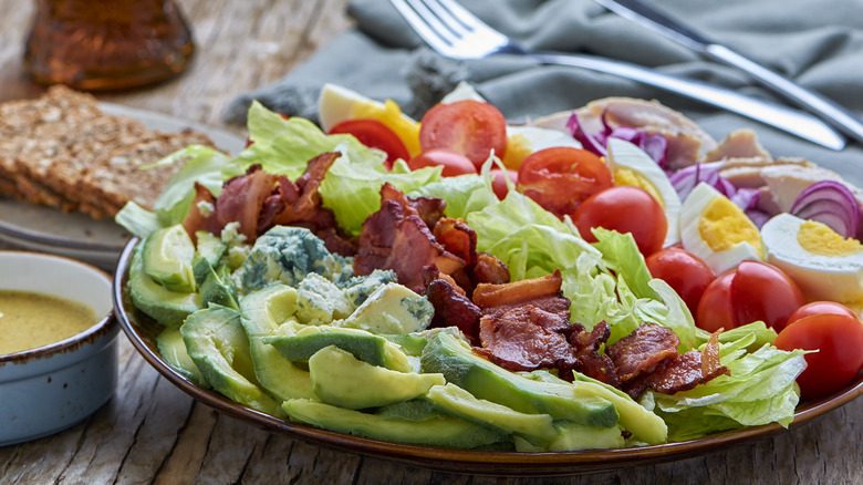 A Cobb salad with bacon, avocado, blue cheese, lettuce, hardboiled eggs, tomatoes, and onions on a plate next to a container of dressing
