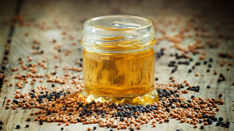 Glass jar of mustard oil surrounded by yellow and black seeds