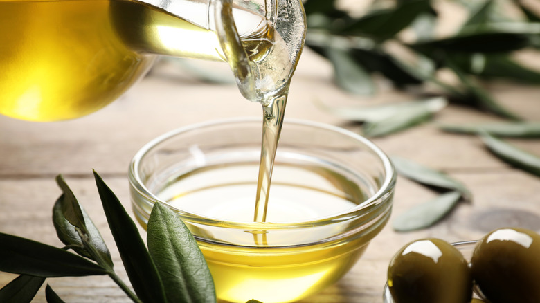 Olive oil being poured into bowl surrounded by olives and leaves