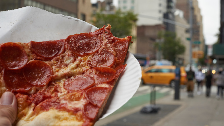 Person holding pizza slice on a paper plate on a NYC street