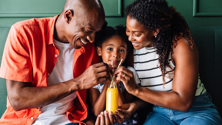 Family with young daughter sharing a glass bottle of orange juice with straws