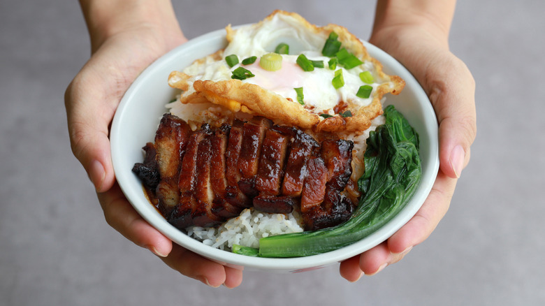 Hands holding bowl with char siu, egg, and greens