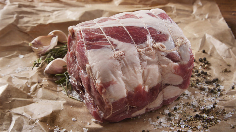 uncooked pork shoulder on butcher paper surrounded by garlic, rosemary, salt, and pepper