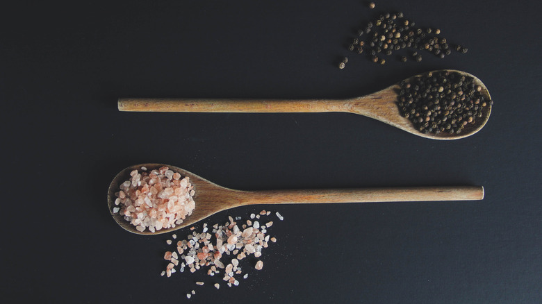 Two wooden spoons holding whole peppercorns and pink salt