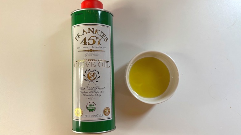 Bottle of Frankies 457 olive oil next to white dish of oil on white table