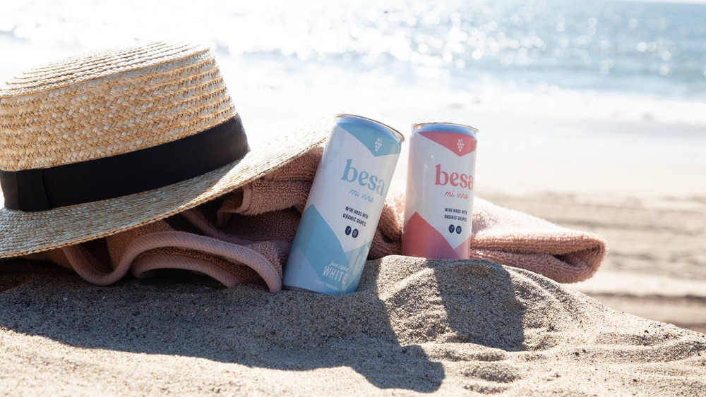 Two cans of wine in the sand at the beach with a towel and straw hat