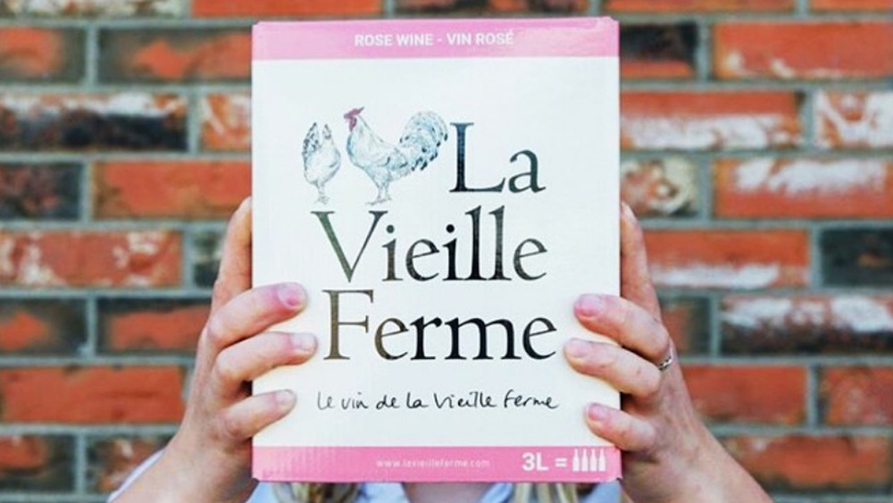 two hands holding a box of la vieille ferme boxed wine in front of a brick wall