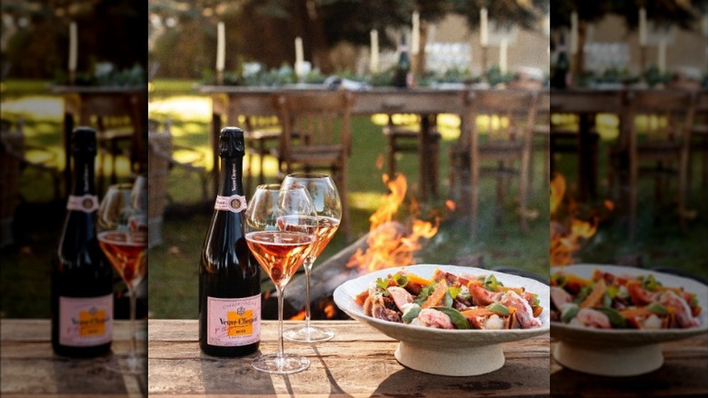 A bottle of rose champagne on an outdoor table with two glasses of wine and a plate of food