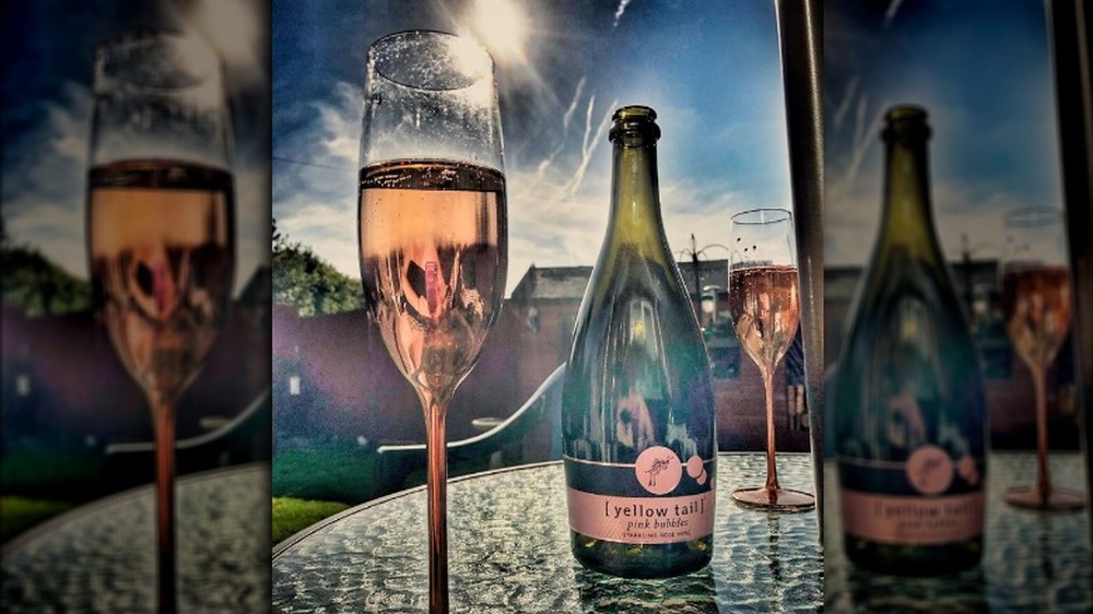 A glass of sparkling rose wine sitting on a glass outdoor table next to the bottle of wine