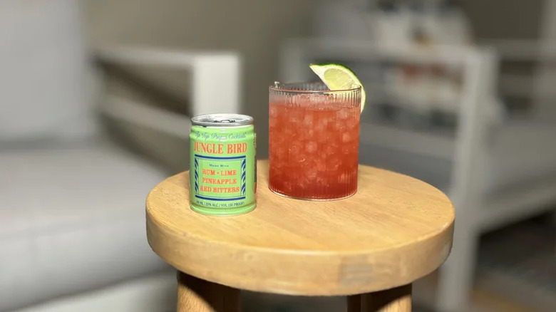 A can of Tip Top Proper Cocktails Jungle Bird next to a glass filled with ice, the drink, and a lime wedge