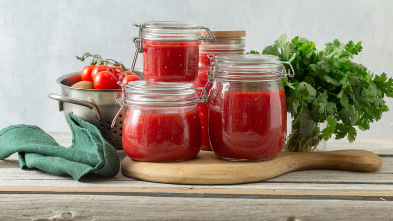 Tomato sauce in glass jars with fresh tomatoes and herbs behind them.