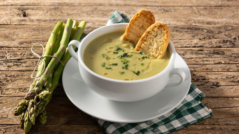 A bowl of creamy asparagus soup next to asparagus spears