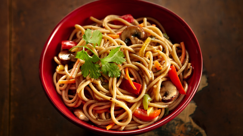 Cooked noodles with vegetables in a red bowl on wood surface