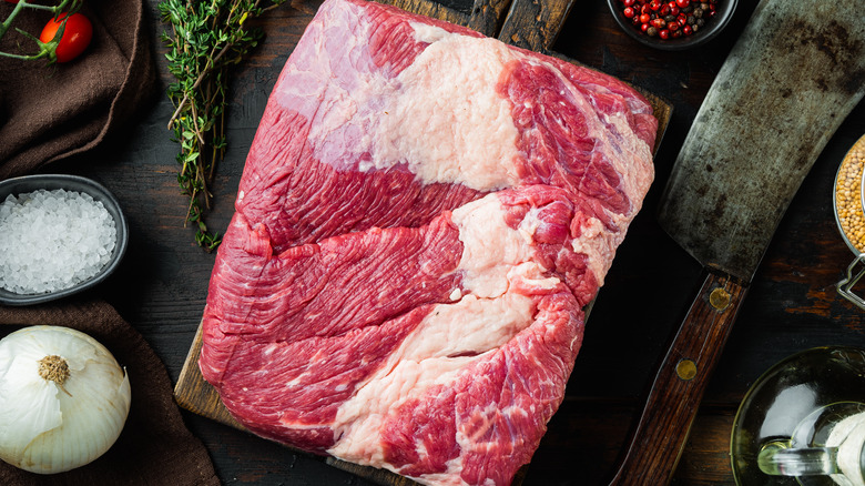 Raw brisket on a cutting board surrounded by a butcher's knife, onion, salt, spices, and herbs.