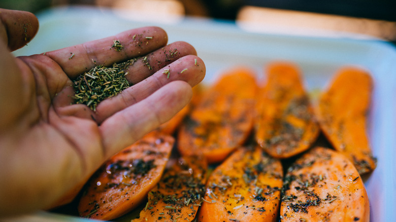 hand holding crushed rosemary over baking dish of sweet potatoes