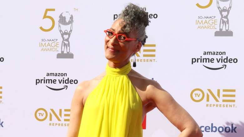 Carla Hall smiling with red glasses and a yellow dress at an event