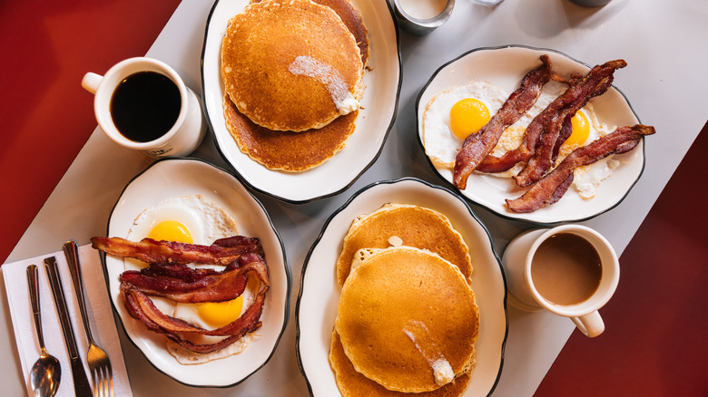 Eggs, bacon, pancakes, and mugs of coffee sit on a table.