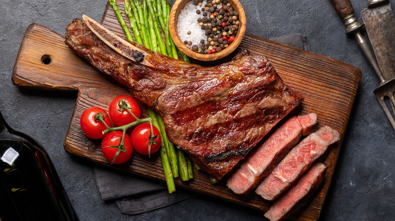 A ribeye steak cut on a wood board with tomatoes, asparagus, and seasonings