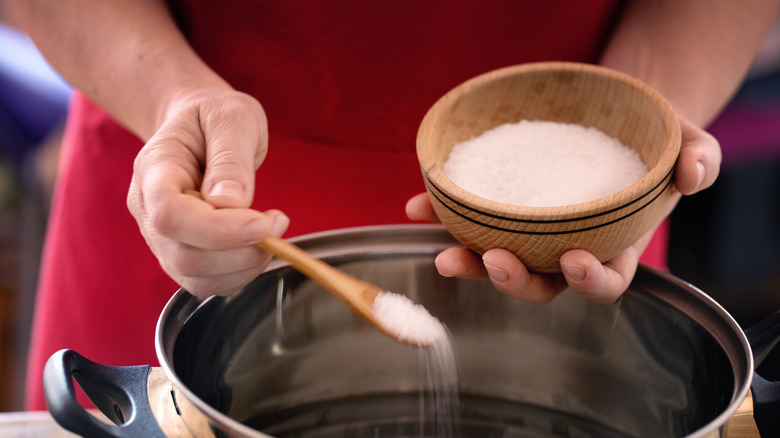 hands adding salt from a wooden bowl to a pot of water