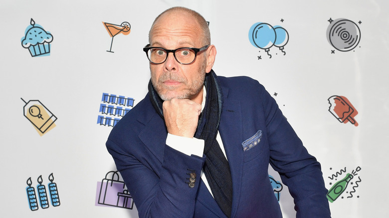 Alton Brown with hand on chin in bue blazer and white shirt, scarf and glasses
