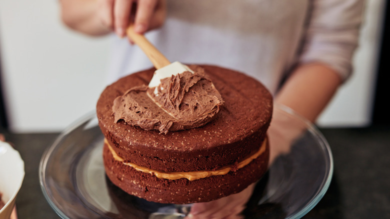 A woman spreading chocolate buttercream onto a layered cake.