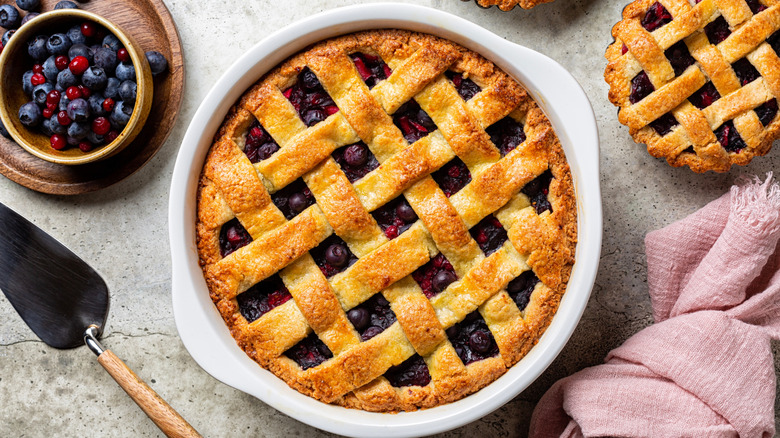 A mixed berry pie in a dish next to a bowl of berries and a mini mixed berry pie.