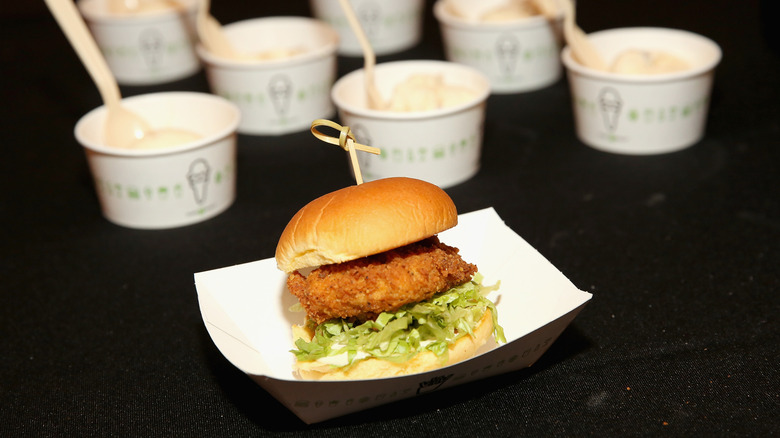 A Chicken Shack sandwich in a paper container with cups of Shake Shack ice cream in the background