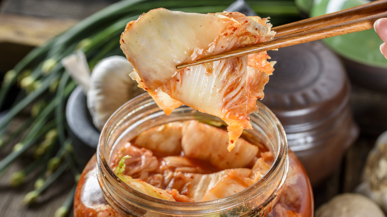 A piece of kimchi held with chopsticks over a jar