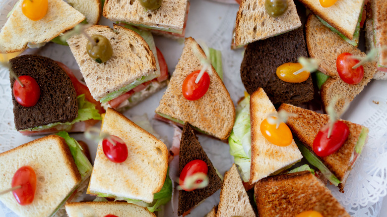 A tray full of small-bite sandwiches with white, wheat and rye bread, skewered with cherry tomatoes on toothpicks